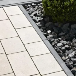 Ułożona ścieżka z jasnych płyt chodnikowych, obramowana szarymi kostkami i kamieniami. W tle widoczne krzewy. Całość tworzy estetyczny wygląd.