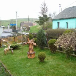 Urokliwy ogród z drewnianym wiatrakiem i figurkami jeleni na tle zielonego trawnika i zabudowań wiejskich. Dekoracyjne krzewy i kwiaty dodają uroku.