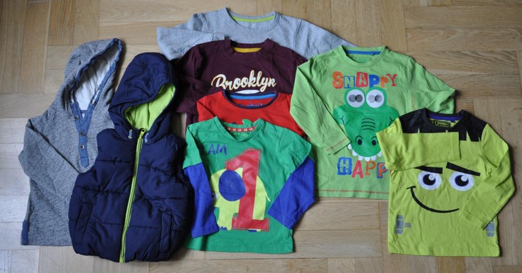 Zestaw używanych ubrań dziecięcych, w tym bluza z kapturem, bezrękawnik i koszulki z długim rękawem z nadrukami zwierząt i napisami, ułożone na drewnianej podłodze.