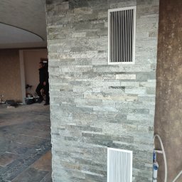 Janusz Zuzelski Uslugi Ogolnobudowlane - Ściana wyłożona kamieniem dekoracyjnym z dwoma kratkami wentylacyjnymi, widoczne fragmenty instalacji hydraulicznej i osoba w tle.