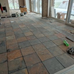 Janusz Zuzelski Uslugi Ogolnobudowlane - Układanie nieregularnych, ciemnych płytek podłogowych w dużej przeszklonej sali, widoczne puszki z klejem, butelka wody i kable.