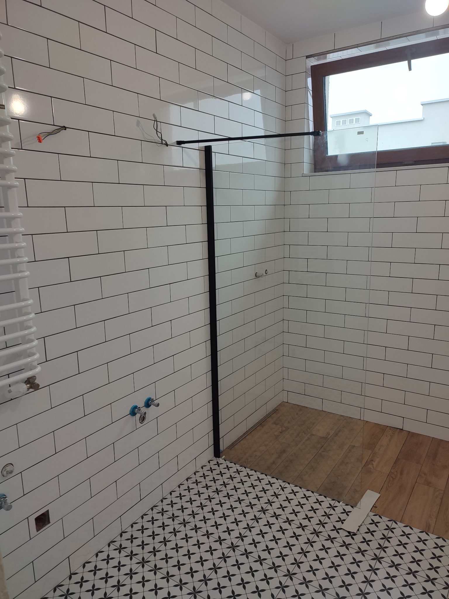 Wnętrze łazienki w trakcie wykańczania: białe, podłużne płytki na ścianach, dekoracyjna podłoga w czarno-białe wzory, fragment drewnianej podłogi w strefie prysznica, widoczne wystające kable i rury.