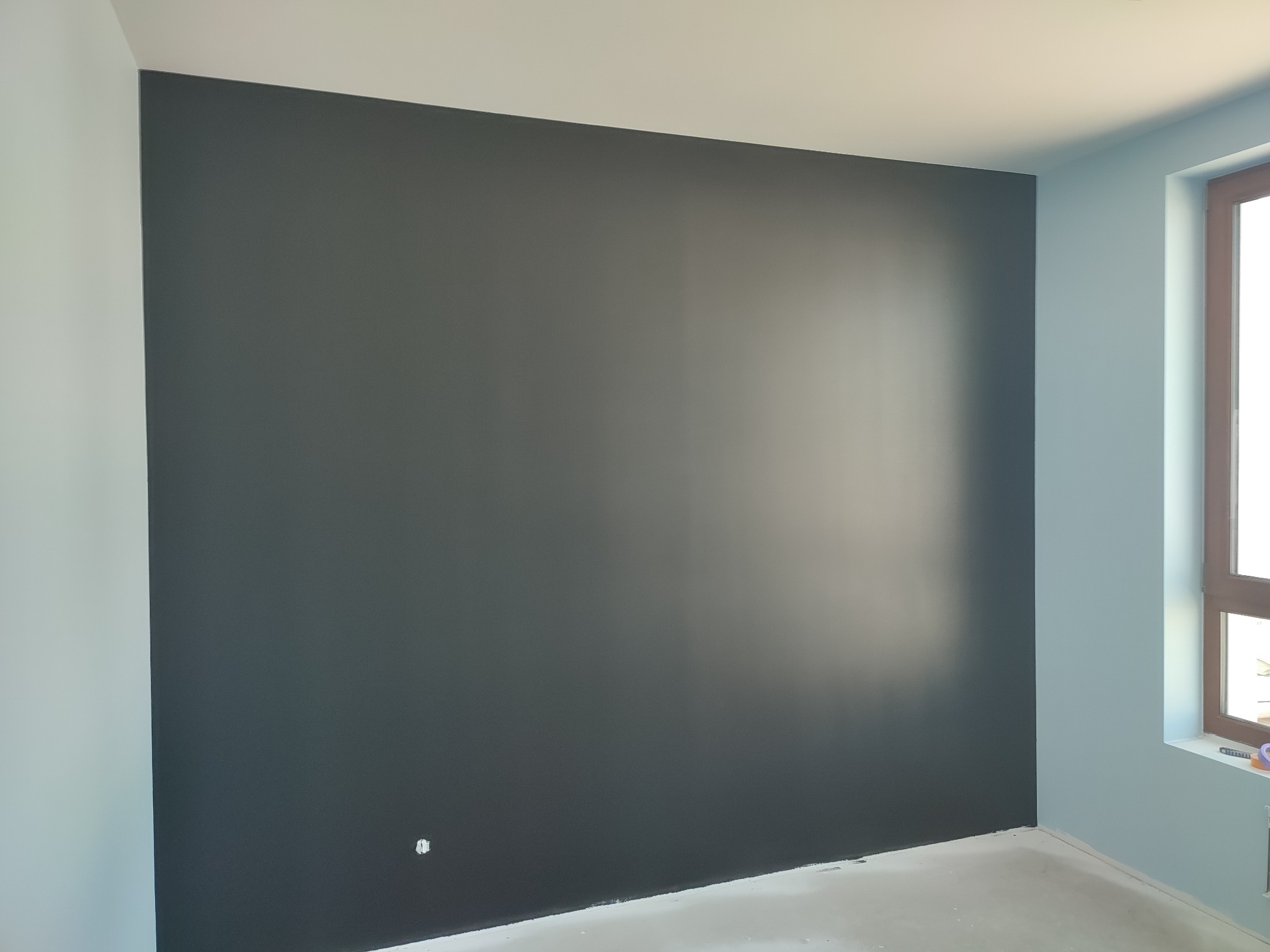 Świeżo pomalowana ściana w odcieniu grafitu, kontrastująca z jasnoniebieskimi ścianami bocznymi i białym sufitem, widoczne gniazdko elektryczne na ciemnej ścianie, surowy stan wykończenia podłogi.