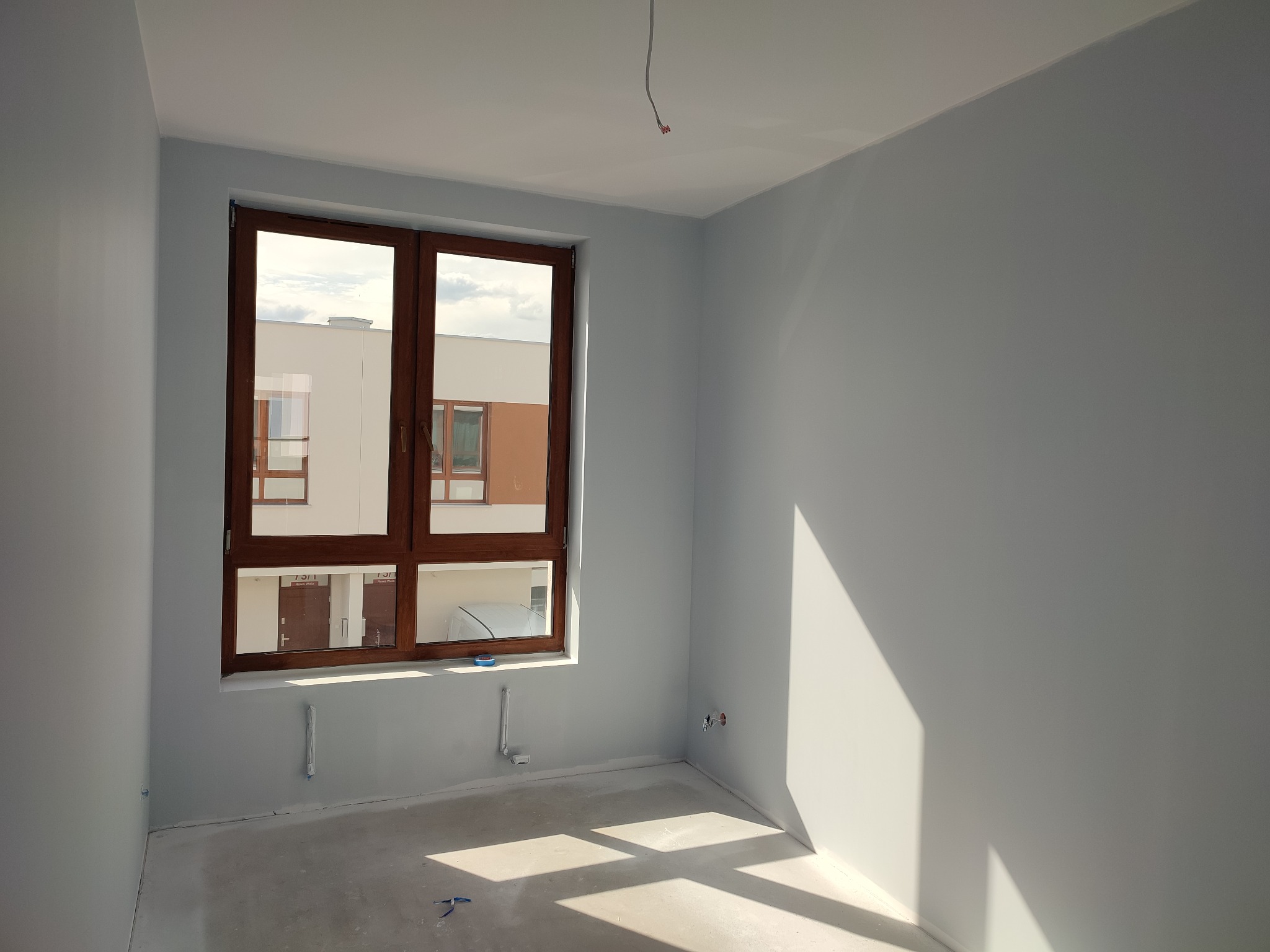 Świeżo pomalowane na szaro ściany w pustym pokoju w Lesznowoli, z widocznymi instalacjami i oknem z brązową ramą, przez które widać budynek mieszkalny.