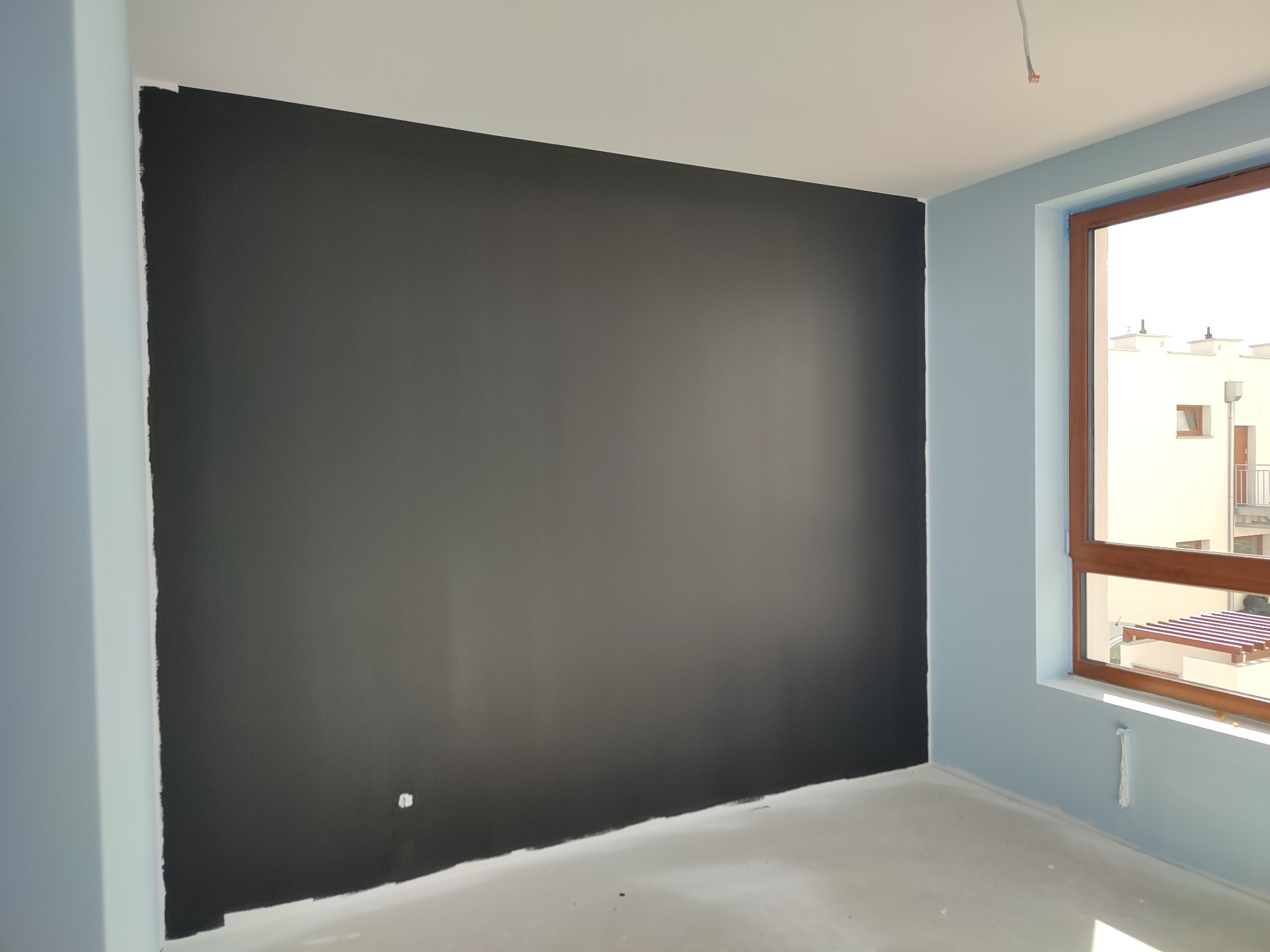 Świeżo pomalowana na czarno ściana w pokoju z niebieskimi ścianami bocznymi i drewnianą ramą okienną, widoczne ślady farby na podłodze i przy suficie.