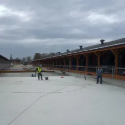 Prace wykończeniowe na betonowej posadzce hali z drewnianą konstrukcją, dwóch pracowników w odblaskowych kamizelkach rozprowadza substancję po powierzchni, widoczne narzędzia pracy i przewody.