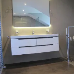 Nowoczesna łazienka z podświetlanym lustrem i białą szafką pod umywalką. Szare płytki na ścianach i podłodze. Widoczny fragment kabiny prysznicowej.