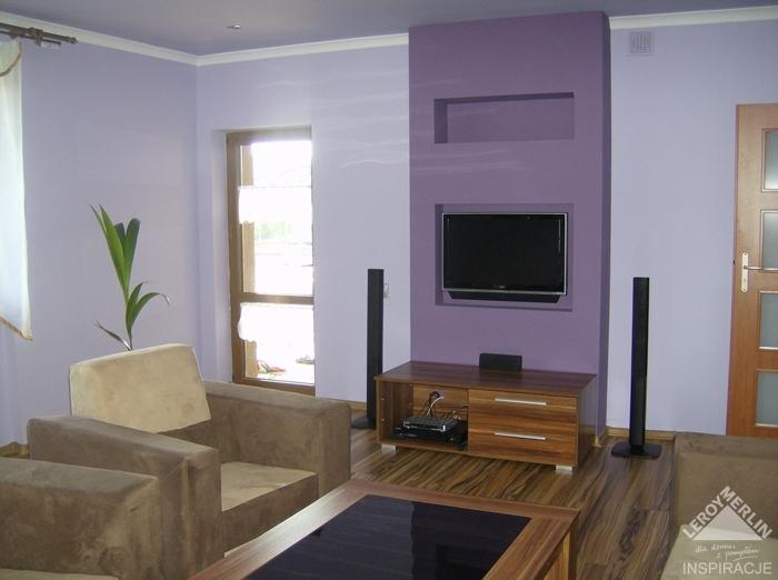 Nowoczesny salon z fioletową ścianą akcentującą, na której wisi telewizor. Widoczne meble w odcieniach brązu i beżu oraz drewniana podłoga.
