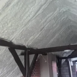 Poddasze po izolacji natryskowej pianką, widoczne krokwie i fragment ściany z cegły, logo firmy PBK w lewym górnym rogu.