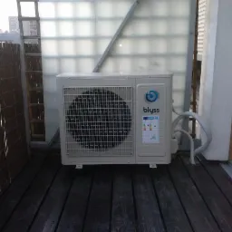 Kompaktowa jednostka klimatyzacji Blyss na balkonie w Trzebini. Widoczne przewody i kratka wentylacyjna. Tło stanowi ścianka z luksferów i drewniana podłoga.