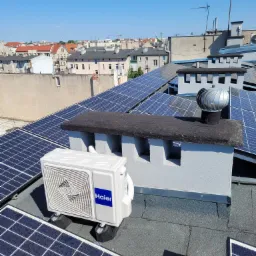 Biała jednostka zewnętrzna klimatyzacji Haier zamontowana na dachu budynku w Sicienko, obok paneli słonecznych i komina wentylacyjnego.