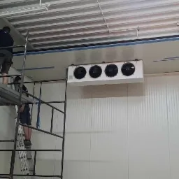 Instalacja chłodnicza z czterema wentylatorami w chłodni, widoczni dwaj monterzy na rusztowaniu i drabinie, białe panele ścienne.