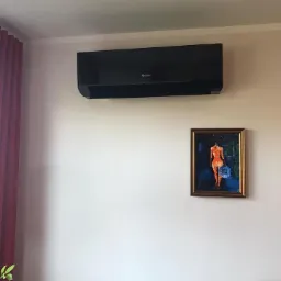 Czarny klimatyzator marki Gree zamontowany na ścianie w pokoju z obrazem i zasłonami.