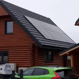 Dom z bali z panelami słonecznymi na dachu, zaparkowany zielony samochód Volvo w tle, pochmurne niebo.