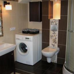 Firma Uslugowa Hydro-Stamar - Ukończona łazienka z pralką, umywalką, toaletą i kabiną prysznicową. Uwagę zwracają brązowe akcenty i dekoracyjne płytki z motywem roślinnym.