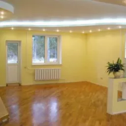 Żółte wnętrze pokoju po remoncie: parkiet, okno z widokiem na las, grzejnik, dekoracyjna ścianka działowa z kwiatami, podwieszany sufit z oświetleniem.