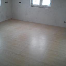 Firma Uslugowa Hydro-Stamar - Jasne, beżowe płytki podłogowe w nowym pomieszczeniu z surowymi ścianami i oknem. Widoczne gniazdka elektryczne, gotowe do wykończenia.