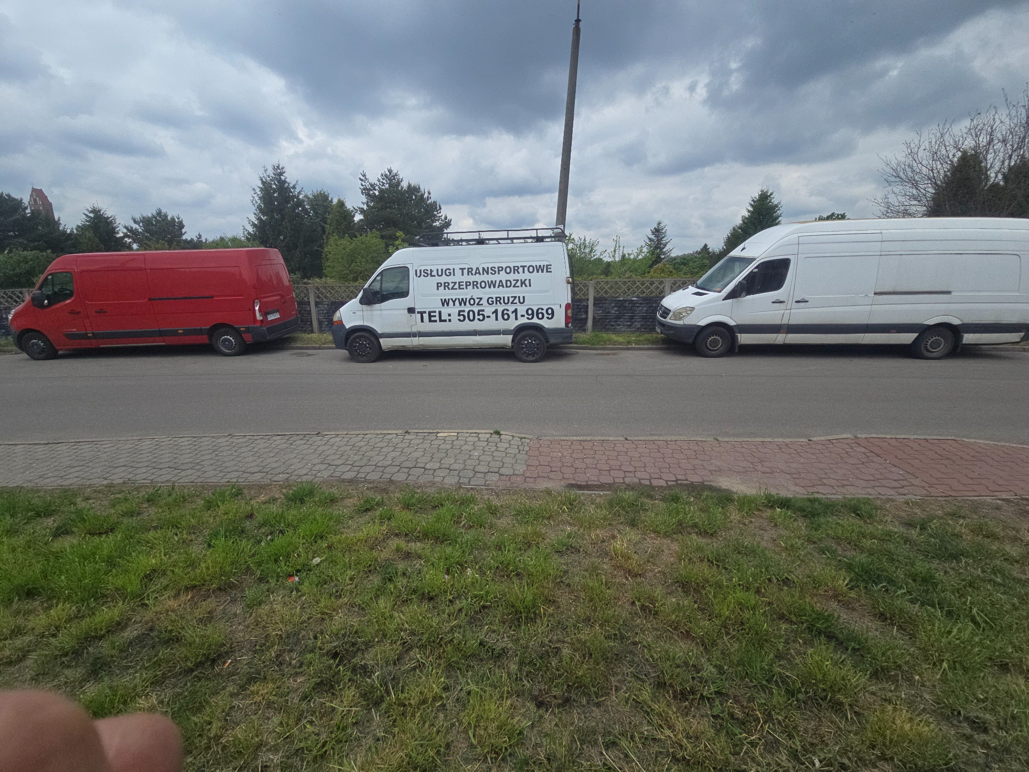 Trzy busy: czerwony, biały z napisem 'Usługi Transportowe Przeprowadzki' i biały. Pojazdy zaparkowane wzdłuż ulicy, widok z boku, pochmurne niebo.