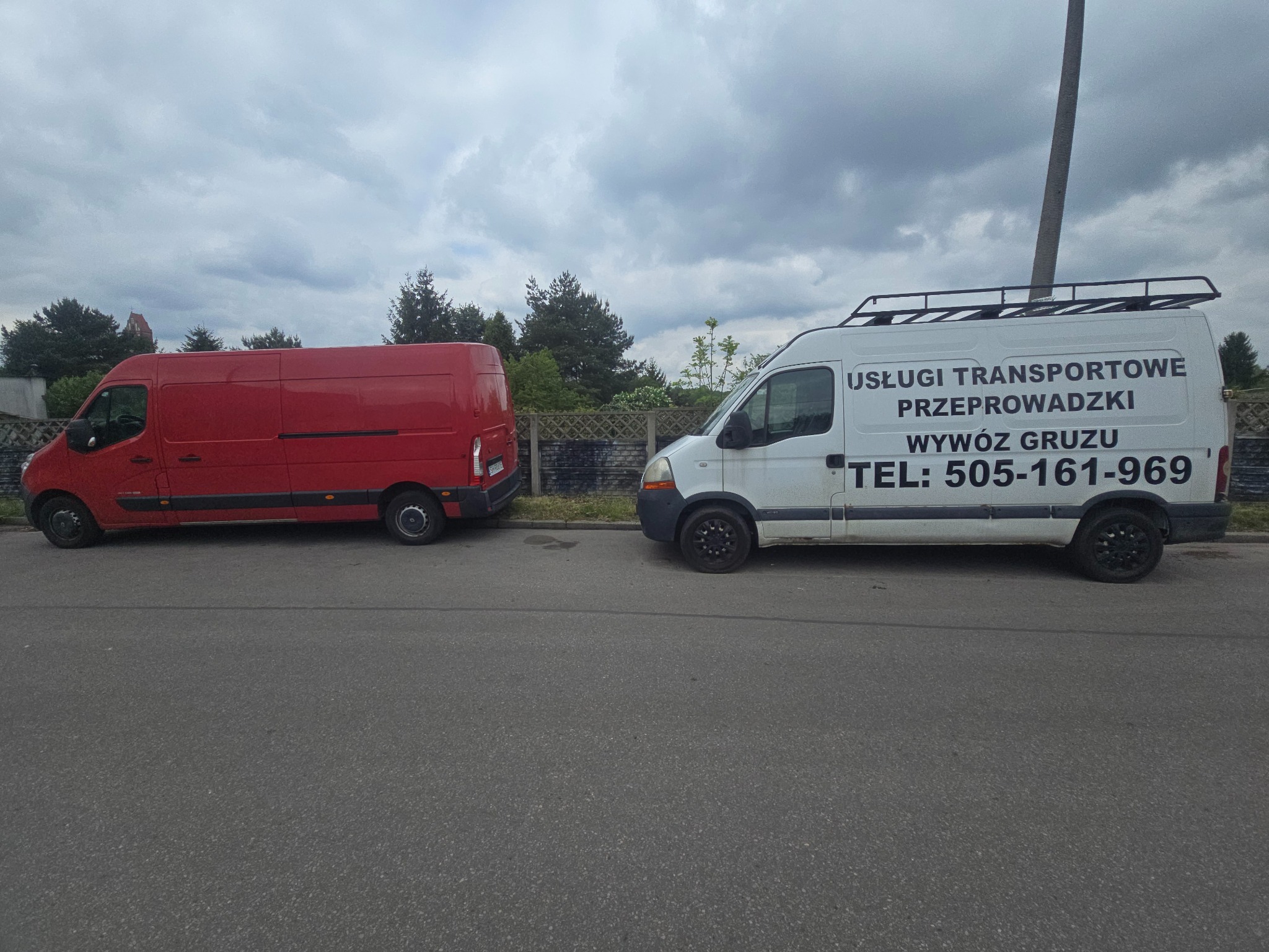 Dwa busy: czerwony i biały z napisem 'Usługi transportowe, Przeprowadzki, Wywóz gruzu' i numerem telefonu, stojące na ulicy w pochmurny dzień.