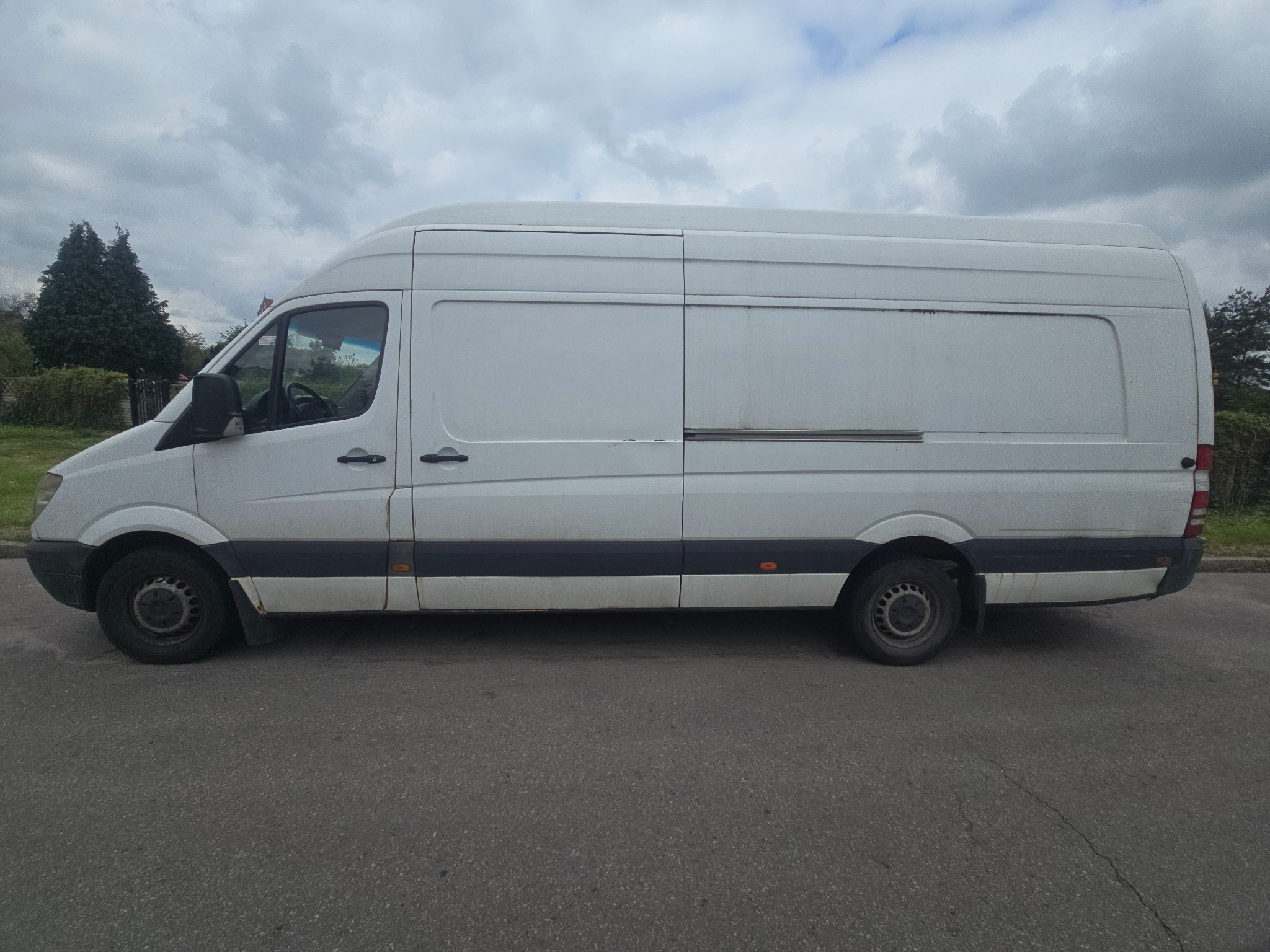 Biały, dostawczy bus Mercedes Sprinter, zaparkowany na asfalcie, widziany z boku na tle pochmurnego nieba. Widoczne detale: relingi dachowe, przesuwne drzwi.
