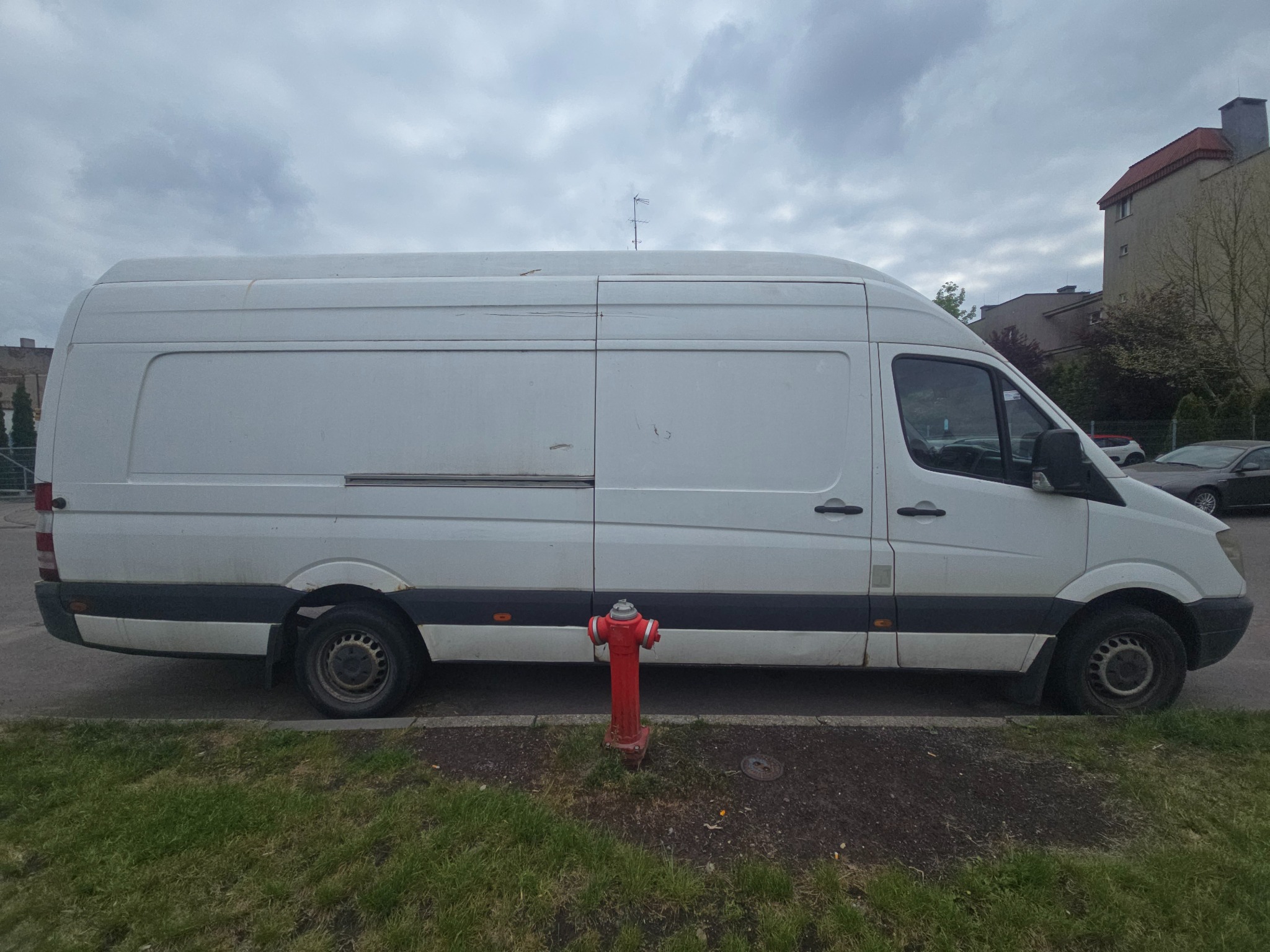 Biały, lekko zabrudzony bus dostawczy zaparkowany na ulicy, z hydrantem pośrodku kadru. W tle budynki i pochmurne niebo.
