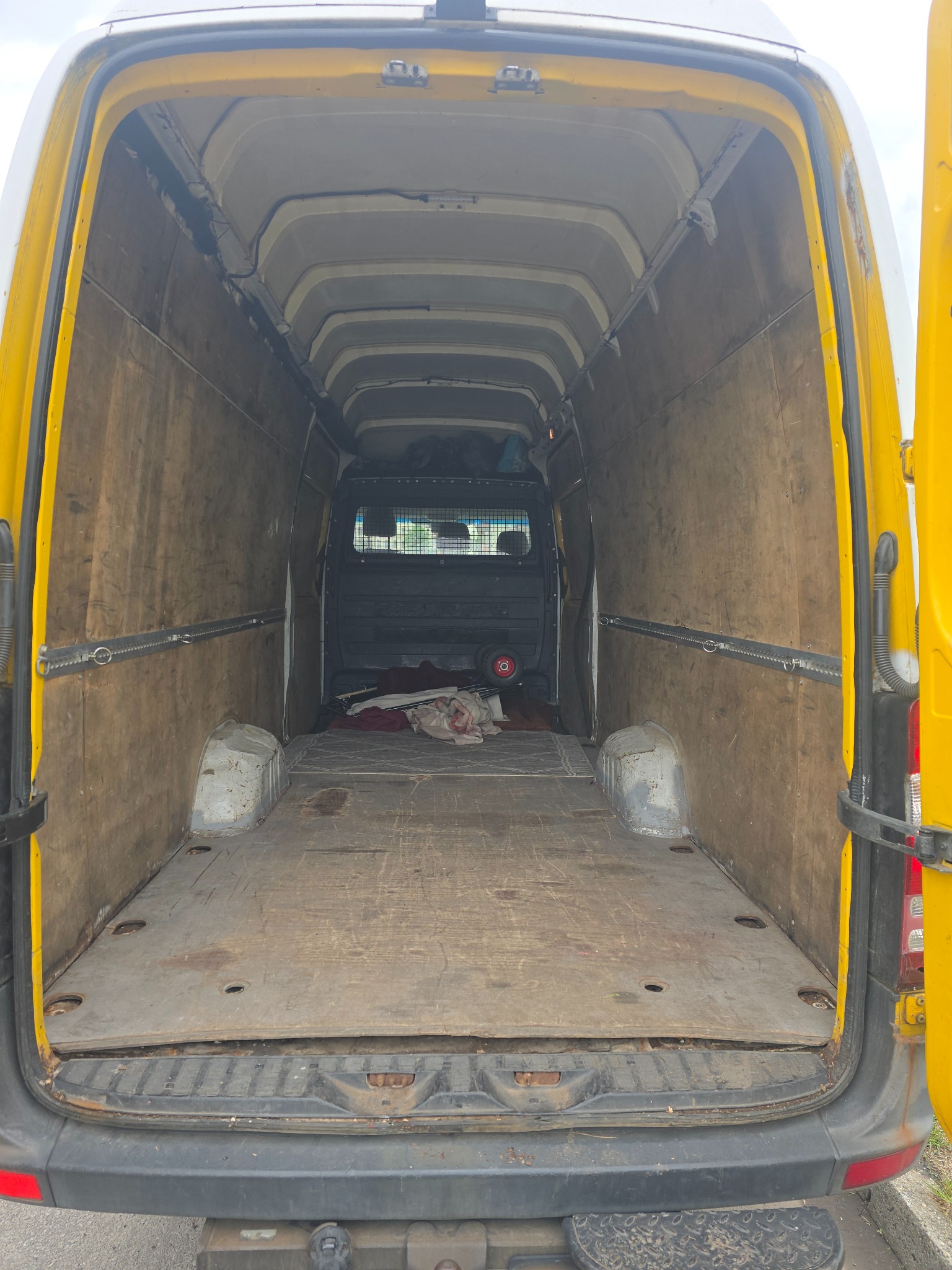 Wnętrze paki żółtego busa transportowego z drewnianą podłogą i ścianami. Widoczne mocowania na ścianach, tył pojazdu z otwartymi drzwiami. Częściowo widoczne przedmioty wewnątrz.