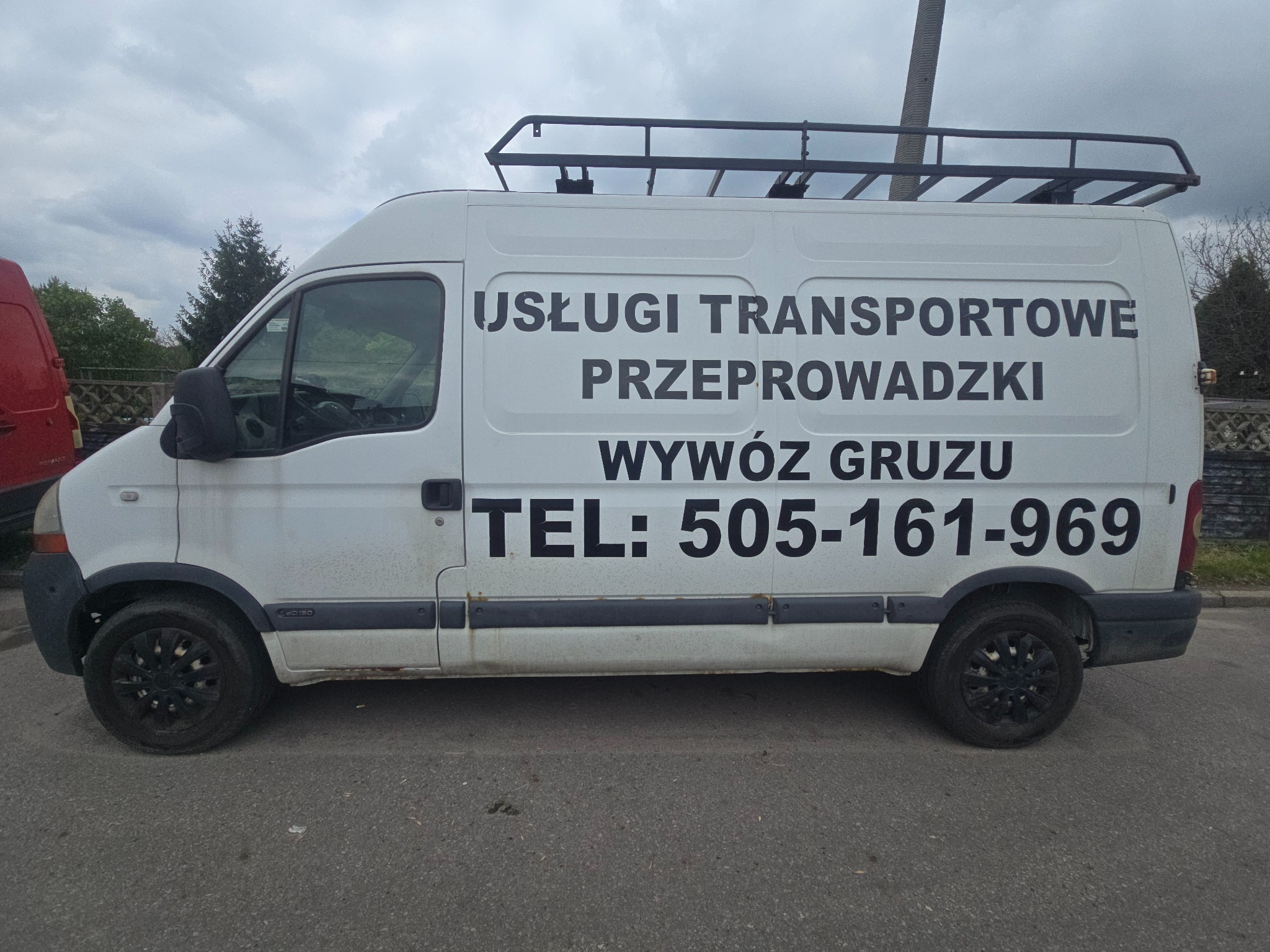 Biały bus z czarnym bagażnikiem dachowym, oferujący usługi transportowe, przeprowadzki i wywóz gruzu, z numerem telefonu na boku, zaparkowany na ulicy.