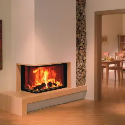 Nowoczesny kominek narożny z paleniskiem, obok pionowy stos drewna. W tle widoczna jadalnia przez otwarte drzwi, jasne wnętrze, narzędzia kominkowe z lewej.