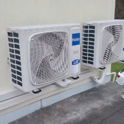 Dwa identyczne, białe klimatyzatory Haier typu split zamontowane na elewacji budynku, widoczne elementy instalacji oraz dwa białe samochody w tle.