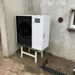 Zewnętrzna jednostka pompy ciepła Mitsubishi Zubadan, zamontowana na białym stelażu przy szarej ścianie budynku z widocznymi śladami wilgoci, obok kostki brukowej i rabaty ziemi z plastikowymi...