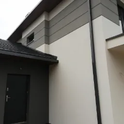 Nowoczesny dom z elewacją w odcieniach szarości i bieli, widoczna boczna ściana z oknami i ciemnymi drzwiami wejściowymi, czarny dach z rynną.