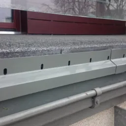 Detale wykończenia balkonu z szarą, metalową balustradą i oknem w tle, widoczne otwory w profilach.