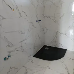 Wykończona łazienka z białymi marmurkowymi płytkami i czarnym brodzikiem, widoczne wystające rurki instalacji hydraulicznej przed montażem armatury.