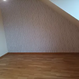 Pomalowane poddasze ze wzorzystą tapetą na skośnej ścianie, widoczna drewniana podłoga i gniazdko elektryczne.