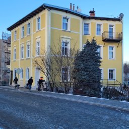 RB - Żółta, odnowiona kamienica z balkonem, widziana z poziomu ulicy wyłożonej kostką. Trzech pracowników przy balustradzie, jeden z taczką. Błękitne niebo w tle.