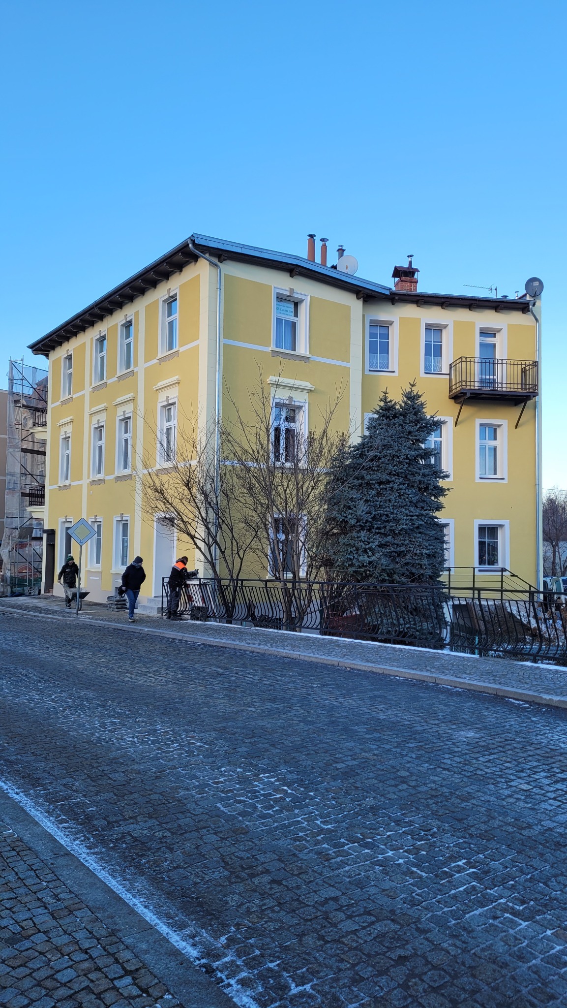 Żółta, odnowiona kamienica z balkonem, widziana z poziomu ulicy wyłożonej kostką. Trzech pracowników przy balustradzie, jeden z taczką. Błękitne niebo w tle.