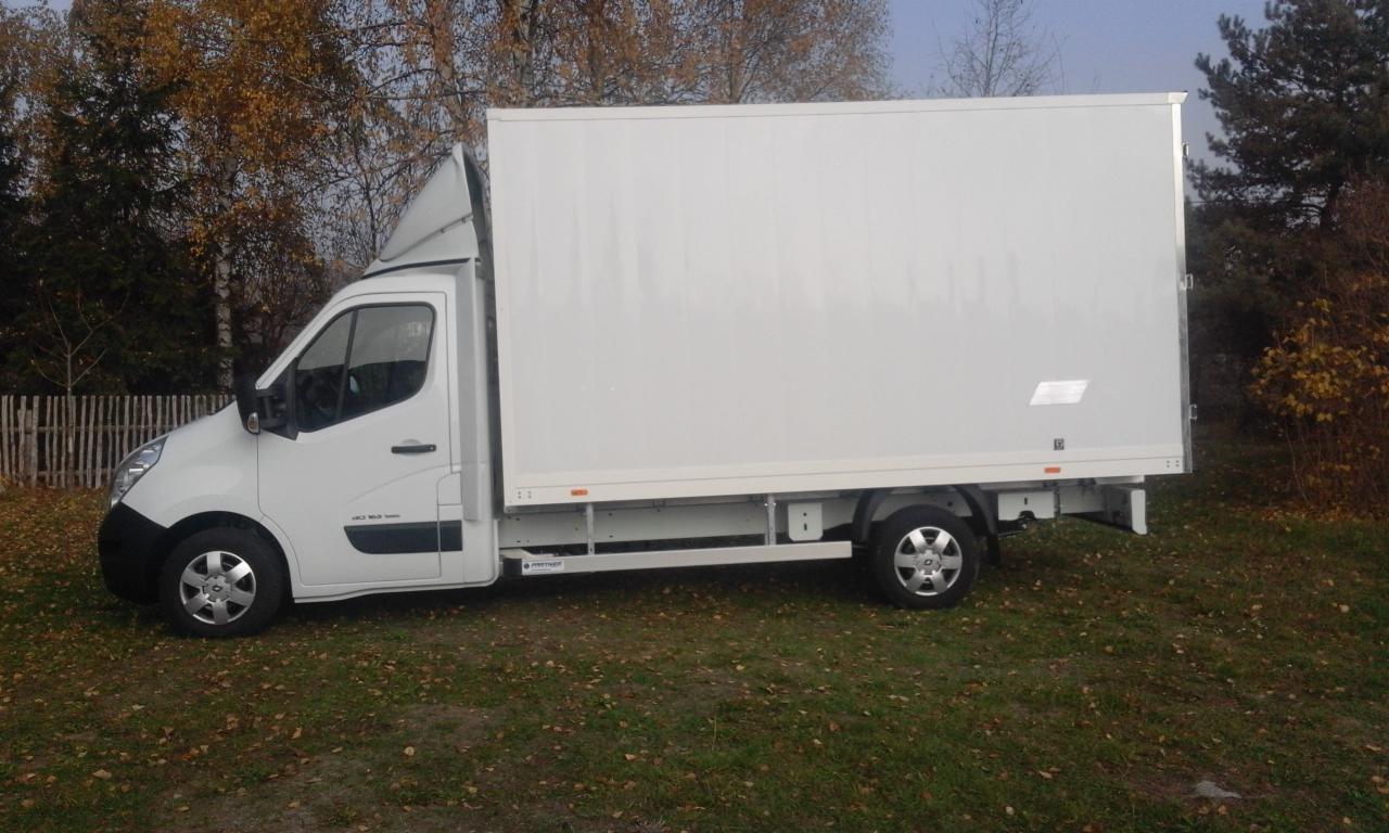 Biały samochód dostawczy z zabudową kontenerową zaparkowany na trawie, widok z boku, jesienne drzewa w tle.