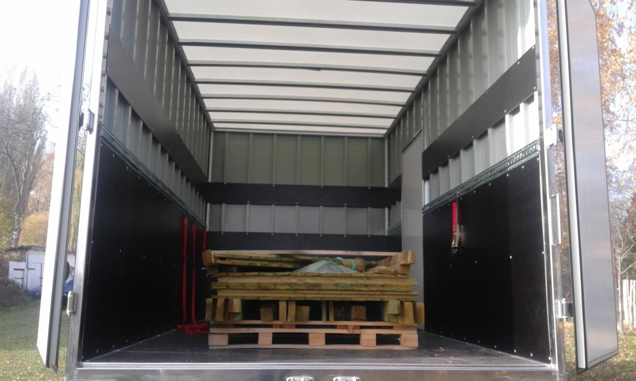 Wnętrze paki transportowej z zabezpieczonym ładunkiem drewnianych elementów na palecie, widoczne pasy mocujące. Otwarta przestrzeń ładunkowa z widokiem na zewnątrz.