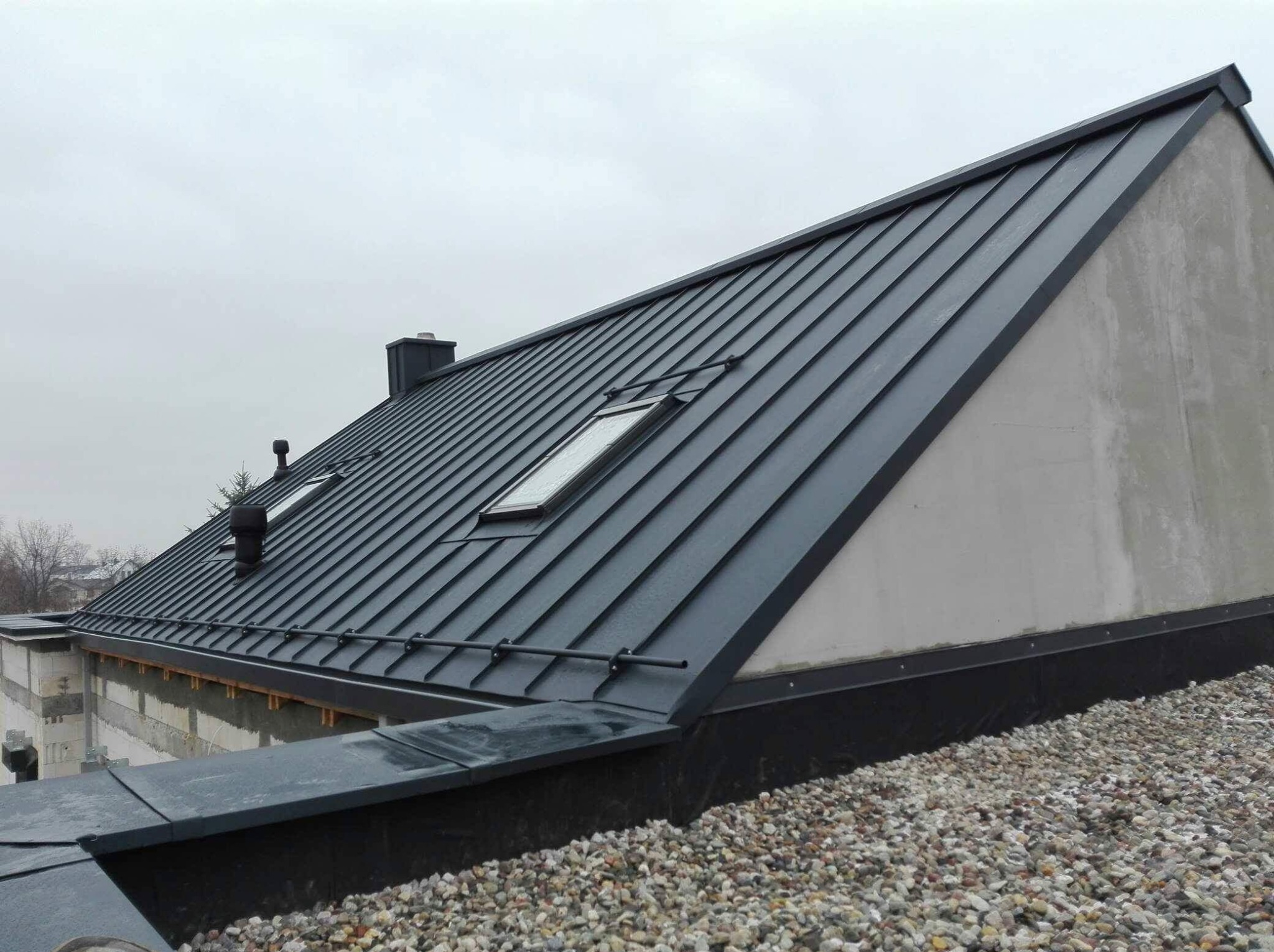 Nowoczesny dach z blachy na budynku z oknami dachowymi i kominami, widoczny z perspektywy dachu pokrytego żwirem, pochmurne niebo w tle.