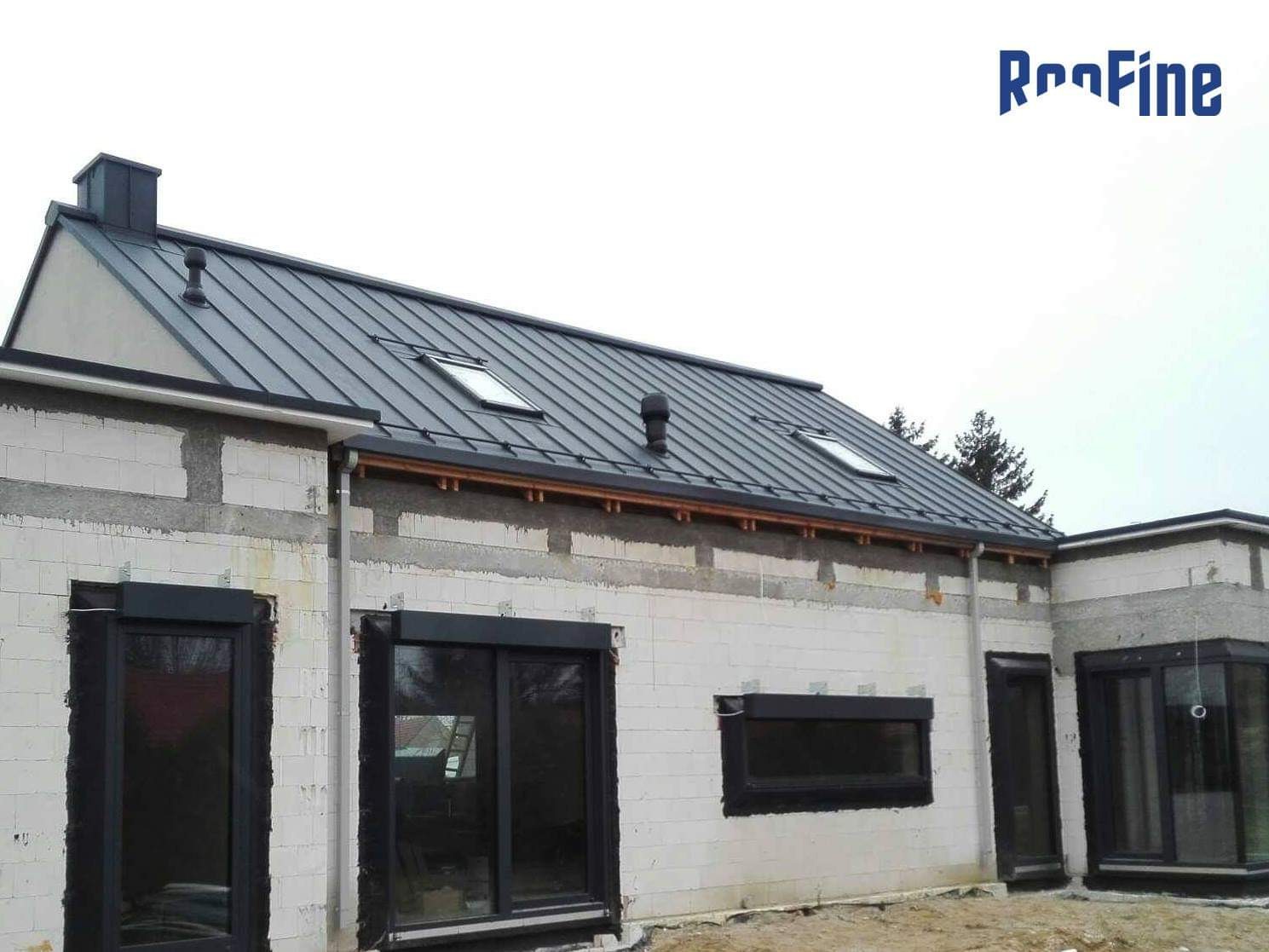 Nowoczesny dom w trakcie budowy z szarym dachem panelowym, widoczne okna dachowe, rynny i obróbki blacharskie, ściany z białego bloczka.