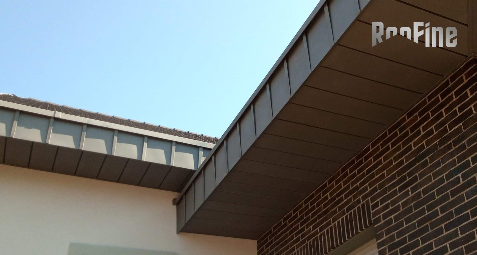 Szczegółowe ujęcie metalowej konstrukcji dachu z widocznym logo producenta 'Roofine' w prawym górnym rogu, na tle błękitnego nieba i ceglanej ściany budynku.