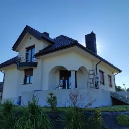 Nowo otynkowany dom jednorodzinny z ciemną dachówką, widoczna drabina i pracownik na elewacji, świeżo zagospodarowany teren wokół budynku.