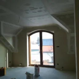Poddasze po szpachlowaniu, widoczne płyty gipsowe na suficie i ścianach, przygotowane do dalszych prac wykończeniowych. Okno w drewnianej ramie w tle.
