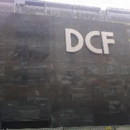 Elewacja budynku wyłożona ciemnym kamieniem z białym logo 'DCF'. Widoczne rusztowania po prawej stronie budynku. Kamienna faktura z wyraźnym wzorem.