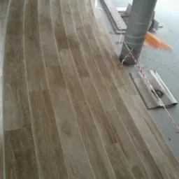 Układanie beżowych płytek imitujących drewno na schodach o zakrzywionym kształcie, z kolumną w tle zabezpieczoną taśmą ostrzegawczą.