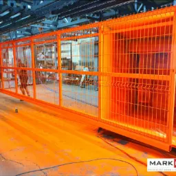Pomarańczowa, metalowa brama przemysłowa w trakcie malowania proszkowego w hali produkcyjnej, widoczne osoby w tle i logo producenta na podłodze.