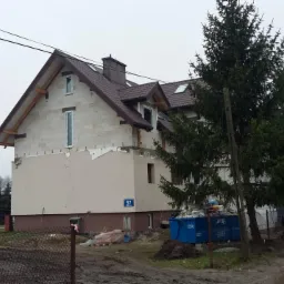 Dom w trakcie budowy z brązowym dachem i widoczną więźbą. Częściowo otynkowana elewacja, okna wstawione. Na pierwszym planie teren budowy i drzewa.