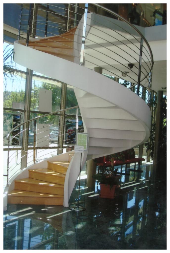 Spiralne schody z drewnianymi stopniami i metalową balustradą w przestronnym wnętrzu z dużymi oknami.