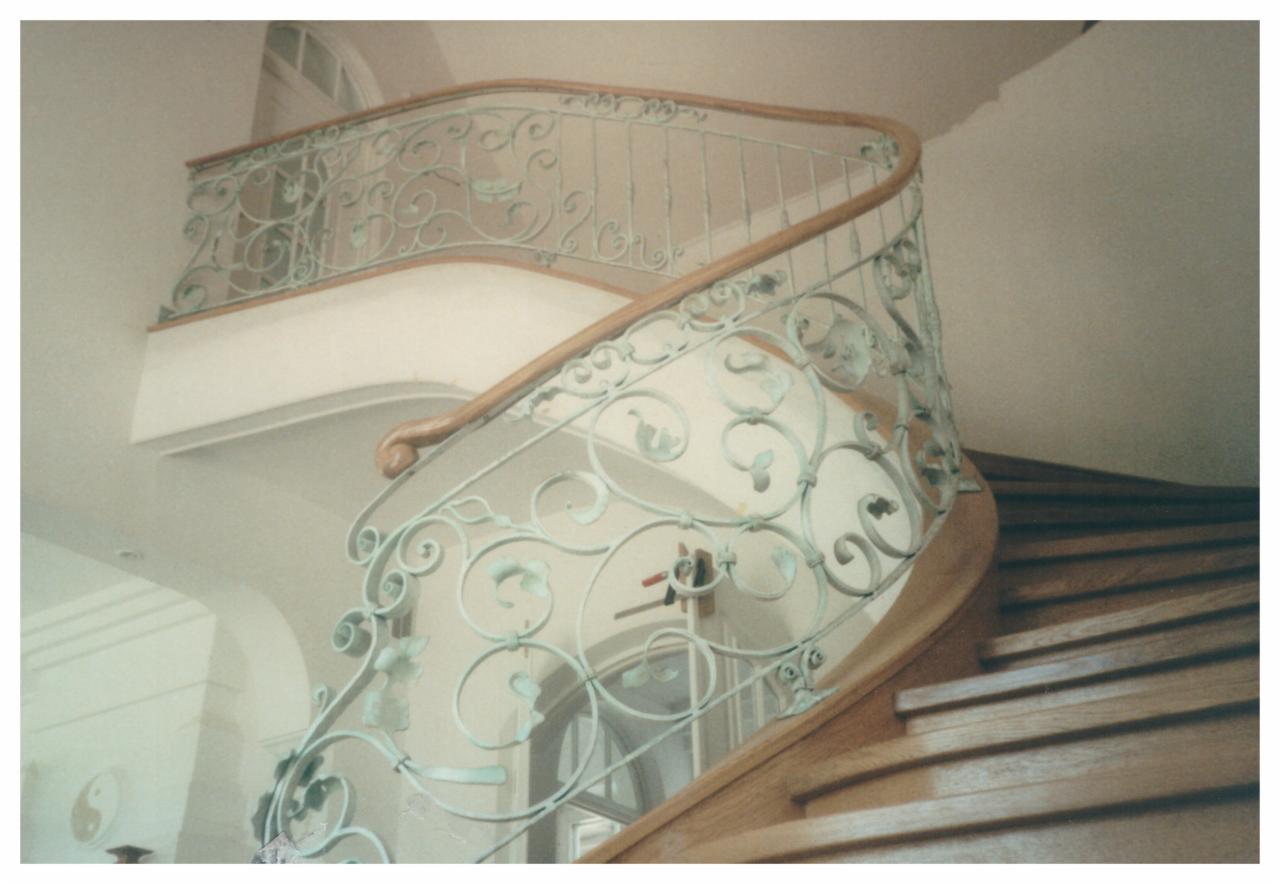 Kręte schody z drewnianymi stopniami i ozdobną, metalową balustradą w kolorze seledynowym z motywami roślinnymi.