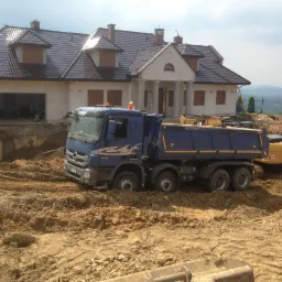 Ciężarówka Mercedes z piaskiem i koparka CAT podczas prac ziemnych przy budowie domu z kolumnami i poddaszem użytkowym.